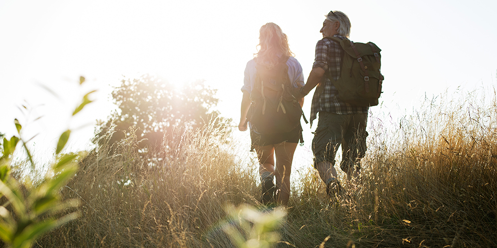 Älterer Mann und ältere Frau beim Wandern über ein Feld im Gegenlicht Älterer Mann und ältere Frau beim Wandern über ein Feld im Gegenlicht